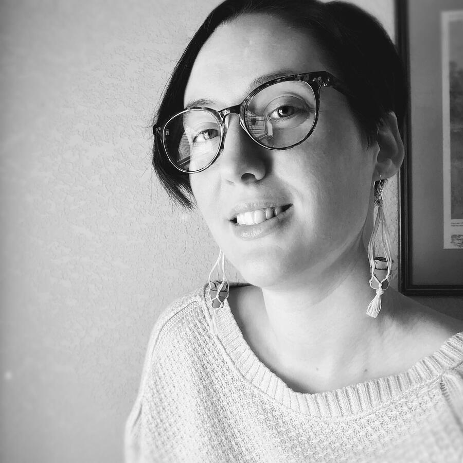 A grayscale photo of the author, Jules Arbeaux, who is wearing glasses and macrame plant earrings.